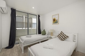 A hotel room with two beds, a window with blinds, a wall-mounted air conditioner, and a framed picture. - Contemporary 2-Bedroom Apartment in Sothbank (South Brisbane)