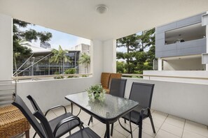A balcony with a glass table and chairs, a small plant, and a view of buildings and trees. - Contemporary 2-Bedroom Apartment in Sothbank (South Brisbane)