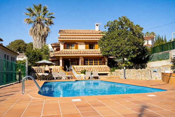 A pool area with a clear blue pool, a palm tree, and a building with a tiled roof.