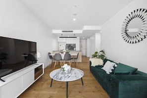 Een moderne woonkamer met een televisie, een ronde salontafel, een groene bank en een eethoek met stoelen, met een keuken op de achtergrond.
