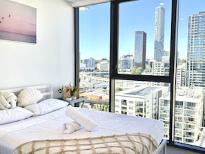 A bedroom with a large window, a bed with white bedding, and a view of a cityscape. - Modern & Vibrant - 1 BR Apt - Pool/Gym/Sauna (South Brisbane)