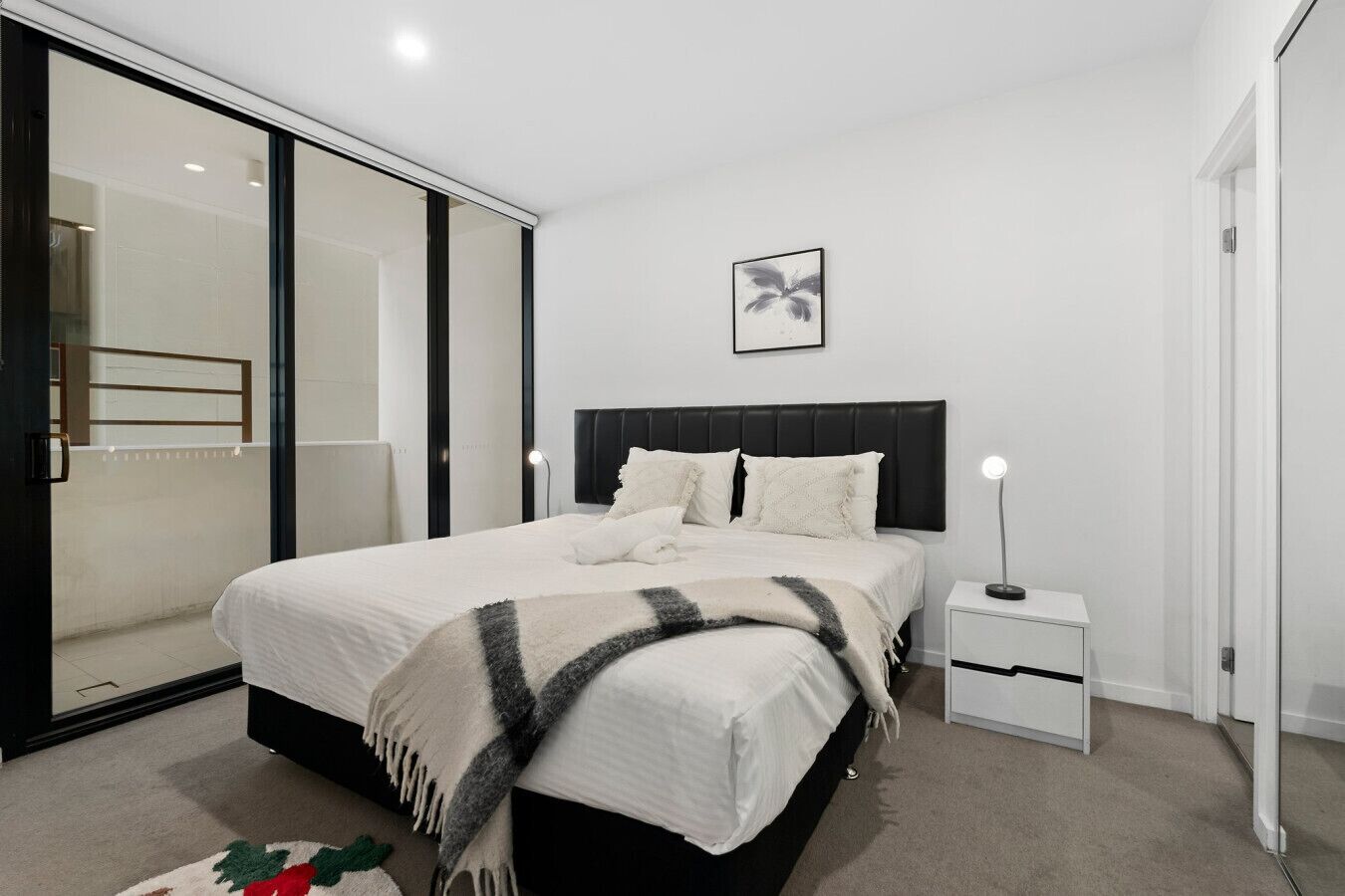 A modern bedroom with a large bed, a bedside table with a lamp, a sliding glass door, and a framed picture on the wall.