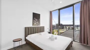Habitación de hotel moderna con una cama grande, una mesita auxiliar pequeña y vistas a la ciudad a través de amplios ventanales.