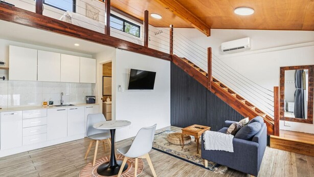Un intérieur moderne comprenant une cuisine, un coin repas et un salon, avec des poutres en bois, un escalier et un téléviseur à écran plat.