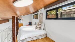 Une chambre avec un plafond en bois, un lit avec une literie blanche, une table de chevet, un ventilateur et une fenêtre donnant sur un paysage verdoyant.
