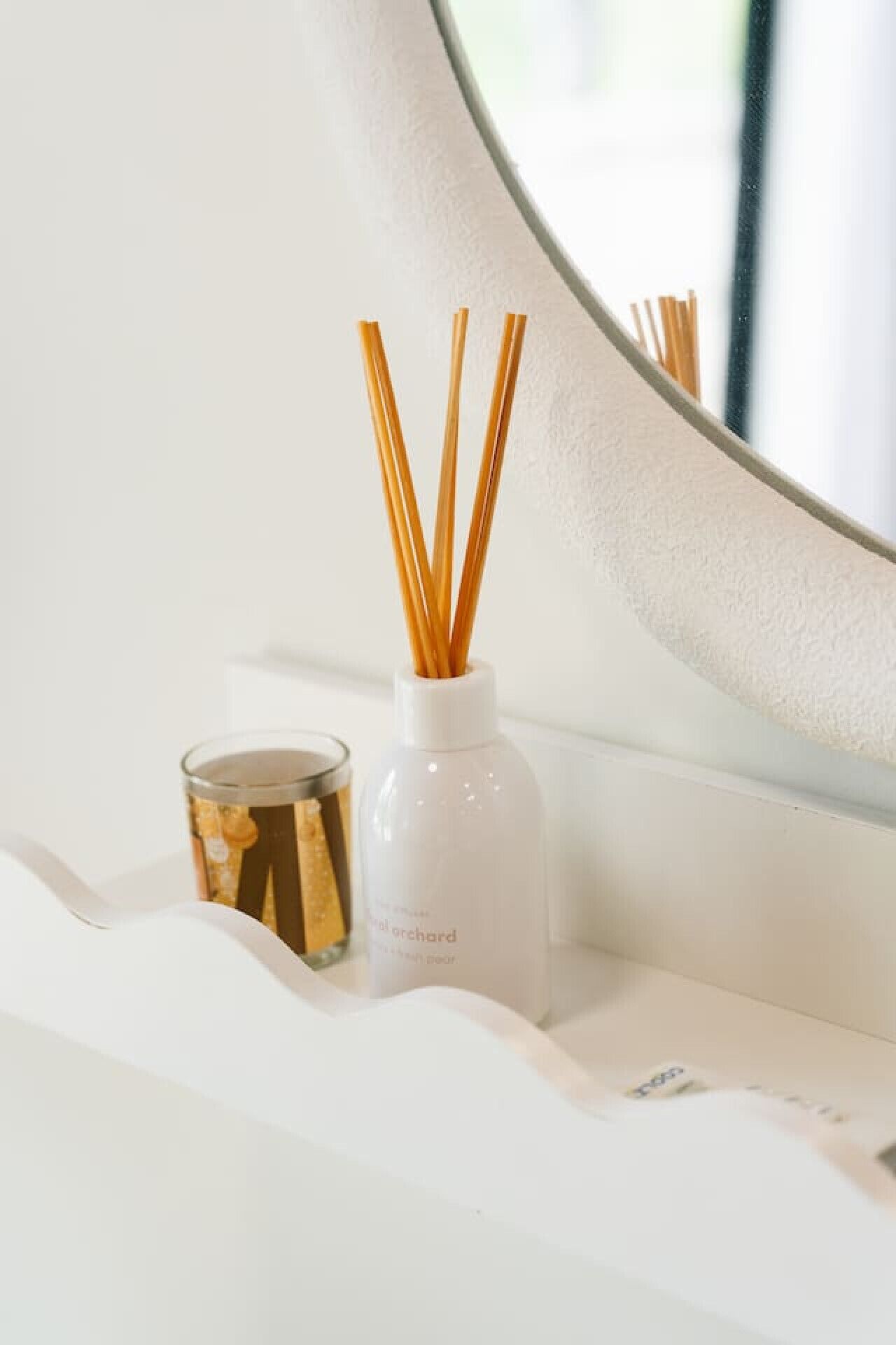 A white bathroom vanity with a round mirror, a white candle, and a reed diffuser.