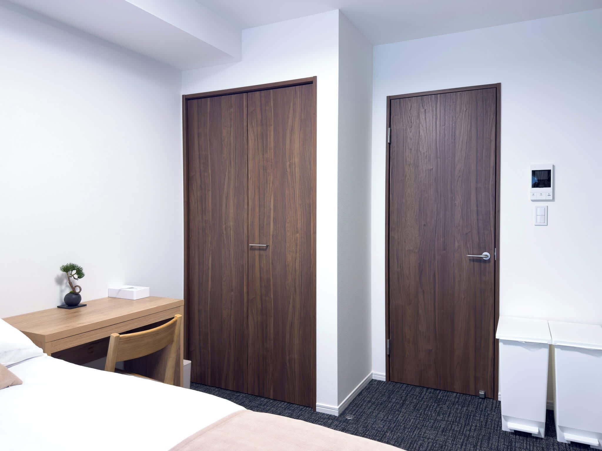 A hotel room with two wooden doors, a desk with a chair, a small plant, and a bedside table.