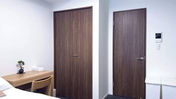 A hotel room with two wooden doors, a desk with a chair, a small plant, and a bedside table.