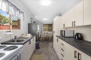 A compact kitchen with a stove, sink, refrigerator, microwave, and a dining area with a table and chairs. - Soothing & Sunny Escape 2B1B @AR2/36 (Acacia Ridge)