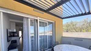 A balcony with a sliding glass door, a table with chairs, and a view of a coastal area. - Perla Blu - Modern Seaside Escape (Preko)