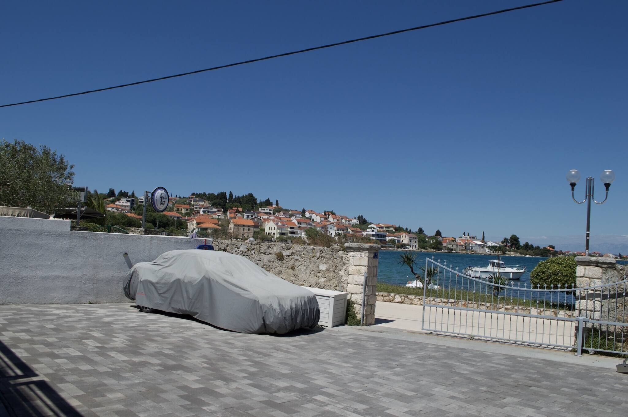 Une voiture à l’abri dans un parking couvert, un mur de pierre, une clôture métallique et une vue sur une ville côtière avec ses bâtiments et ses bateaux.