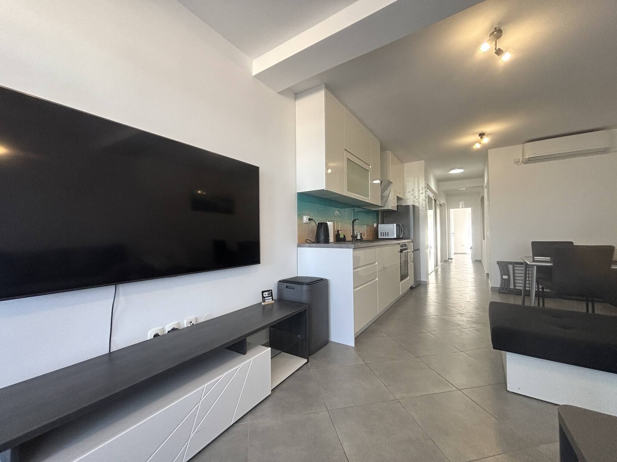 Uma cozinha moderna com televisão de tela plana, uma sala de jantar com mesa e cadeiras, e um corredor que dá acesso a outros quartos.