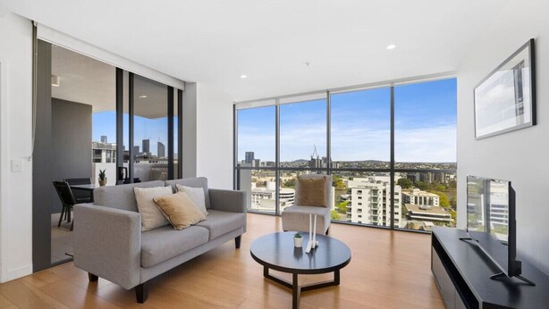 A modern living room with a sofa, armchair, and coffee table, large windows with a city view, and a flat-screen TV on a stand. - Milton apartment-2BR@MT 1905 (Milton)