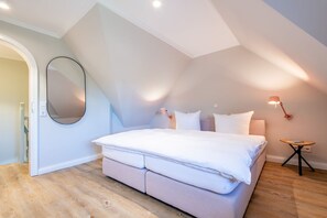 A bedroom with a large bed, a round mirror, a small table, and a wall-mounted lamp. - Friesenhaus Käpt'n Hahn, house part 04 - Friesenhaus Käpt'n Hahn (Sylt)