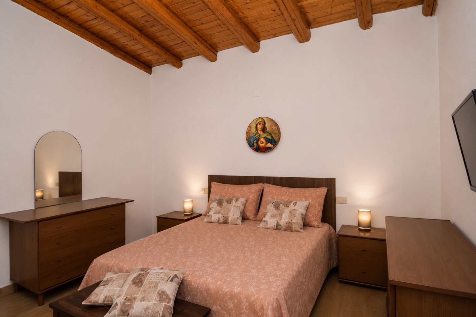 Un dormitorio con techo de madera, una cama con colcha estampada, dos mesitas de noche con lámparas, un tocador y un cuadro colgado en la pared.