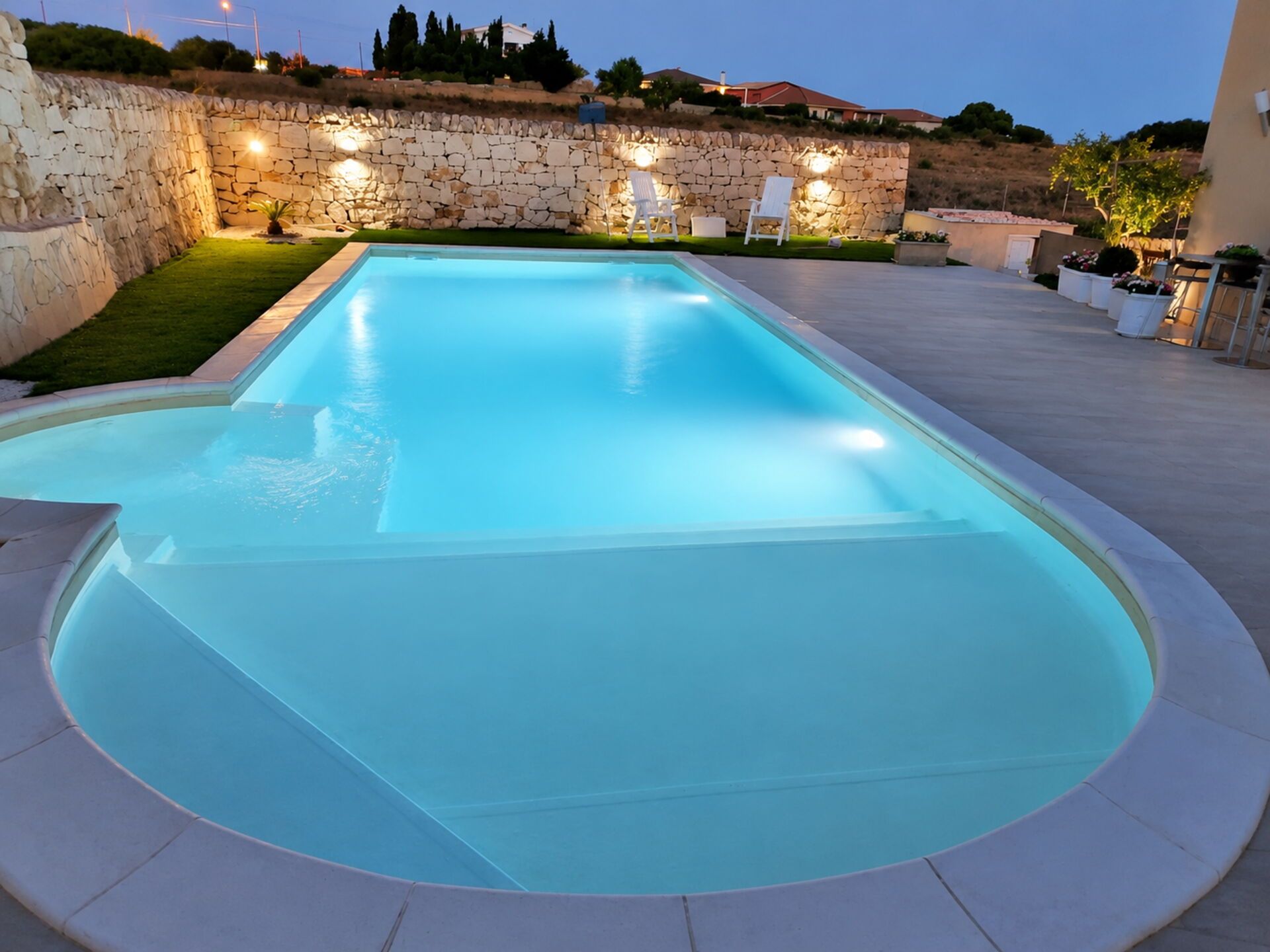 A rectangular swimming pool with a stone wall and lighting, surrounded by a paved area.