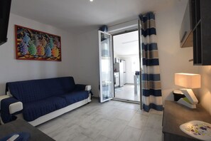 Ein Wohnzimmer mit einem blauen Sofa, einem Gemälde, einer Glastür, die in eine Küche führt, und einer Lampe.
