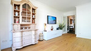 A spacious room with wooden flooring, a large wooden cabinet with glass doors, a white console table, and a wall-mounted TV. - Seesternresidenz Apartment 19 - Seesternresidenz (Scharbeutz)