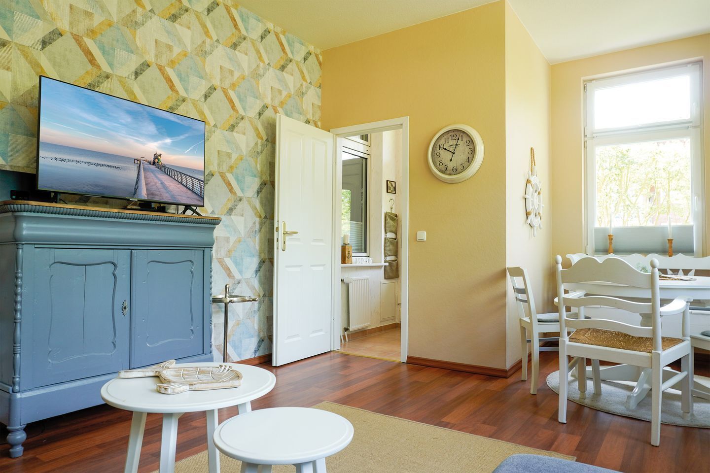 A living room with a television, a dining area with white chairs, a clock on the wall, and a door leading to another room.