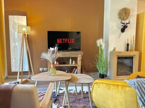 Un salon chaleureux avec une télévision, une cheminée, un canapé jaune et une petite table agrémentée d’objets décoratifs.