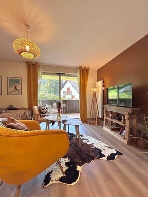 Un salon comprenant un canapé, des fauteuils, une table basse, un téléviseur et une vue sur une maison par la fenêtre.
