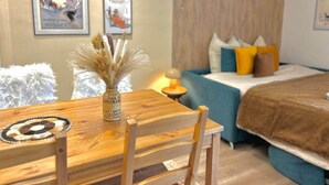 Una habitación acogedora con una mesa de comedor de madera, un jarrón con plantas secas, una cama con ropa de cama azul y marrón, y dos cuadros en la pared.