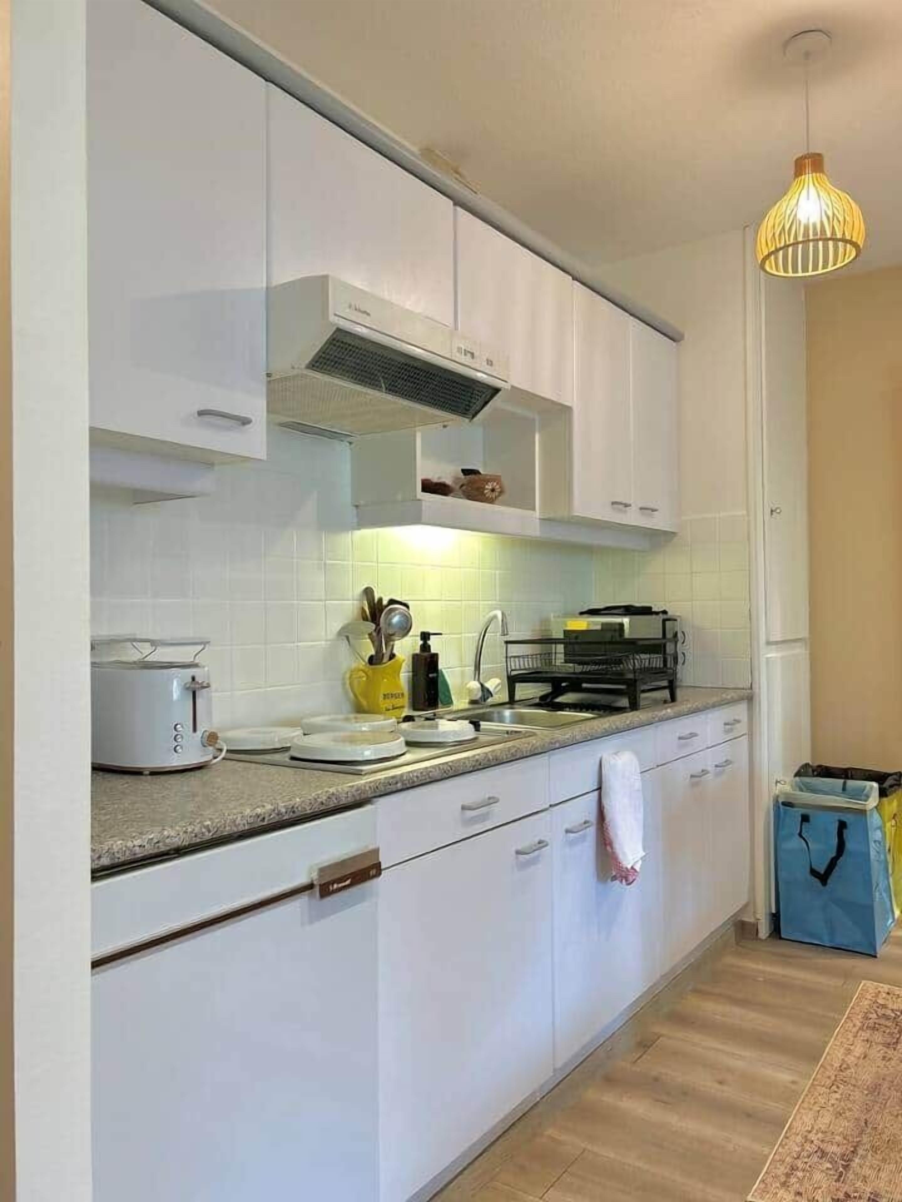 A modern kitchen with white cabinets, a built-in oven, a countertop with a toaster, and a hanging light fixture.
