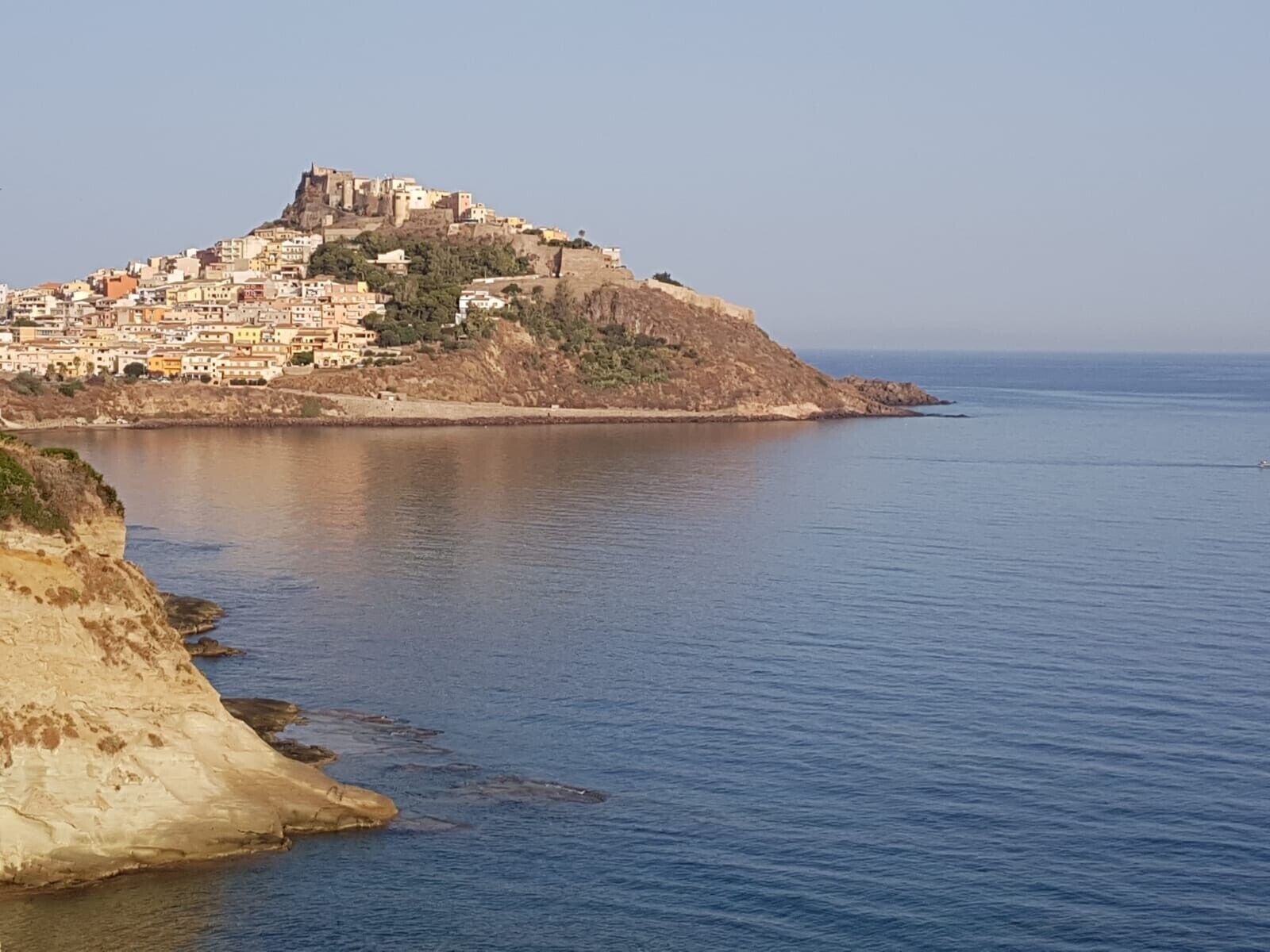 Un pueblo costero con un edificio destacado en la cima de una colina, mar tranquilo y costa rocosa.