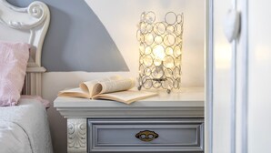Una cama bien hecha con un cojín rosa, una mesita de noche con un libro abierto y una lámpara decorativa.