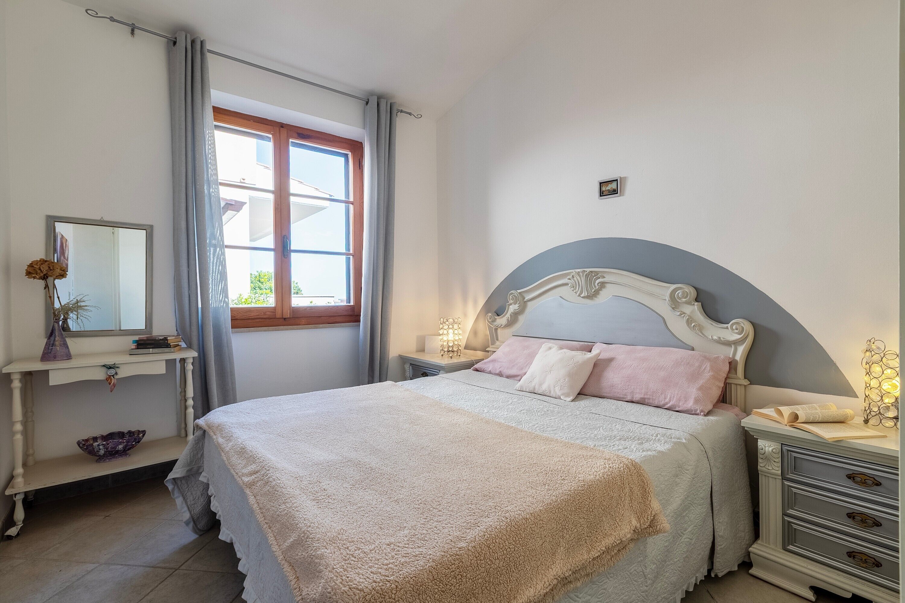 A bedroom with a bed, bedside tables, a nightstand, a mirror, a window with curtains, and a view of the outside.