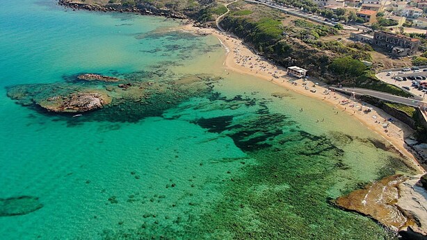 Vista aérea de un pueblo costero con aguas turquesas cristalinas, una playa de arena y un telón de fondo con colinas.