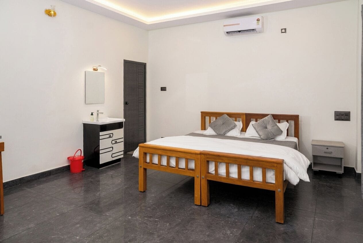 A bedroom with a wooden bed, a bedside table, a nightstand, a red bucket, a white dresser, a mirror, and an air conditioning unit.