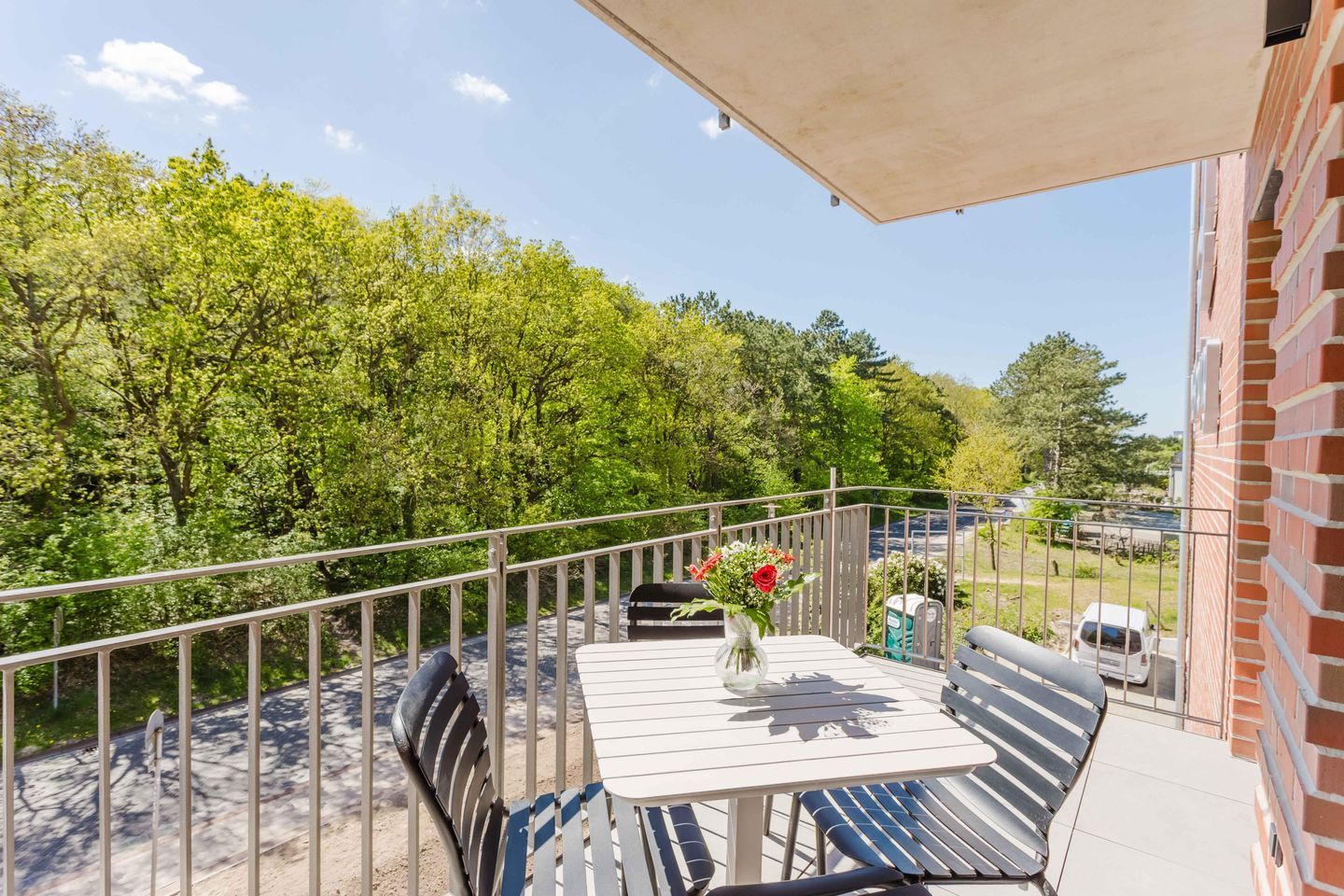 Un balcon avec une table et des chaises, un vase rempli de fleurs et une vue sur des arbres et une route.