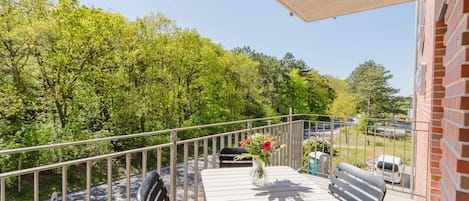 A balcony with a table and chairs, a vase with flowers, and a view of trees and a road.