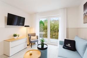 A modern living room with a flat-screen TV, a grey armchair, a sofa, a coffee table, and a balcony with a view of trees. - Haus MEERholung apartment 2.5 - approx. 400 m to the beach for max. 4 guests - (Cuxhaven)