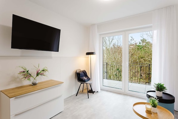 A modern living room with a flat-screen TV, a wooden side table, a chair, and a balcony with a view of trees. - Haus MEERholung apartment 2.6 for up to 5 people, only approx. 400 m to the sea - (Cuxhaven)