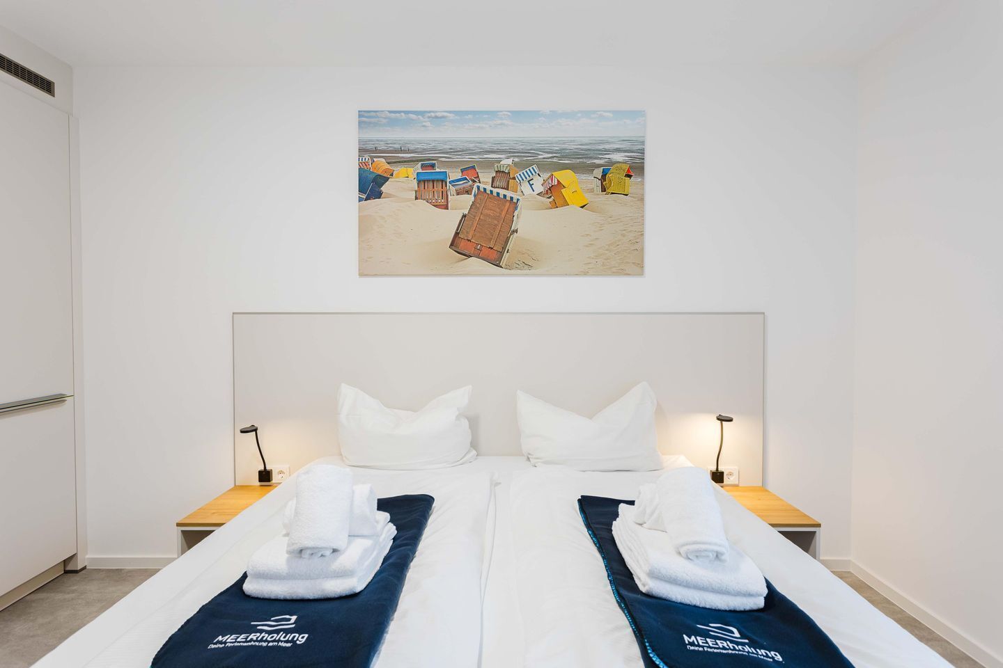 A hotel room with a bed, two bedside tables, a wall-mounted picture of beach chairs, and a white door.