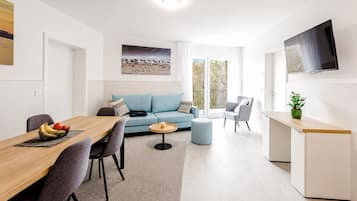 A modern living room with a sofa, a dining table with chairs, a TV mounted on the wall, and a balcony with a view.
