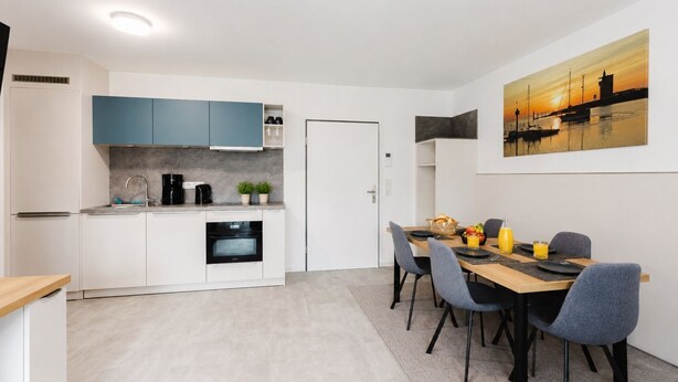 Una cocina moderna con armarios blancos y azules, una zona de comedor con una mesa preparada para dos personas, y un cuadro de un atardecer en la pared.