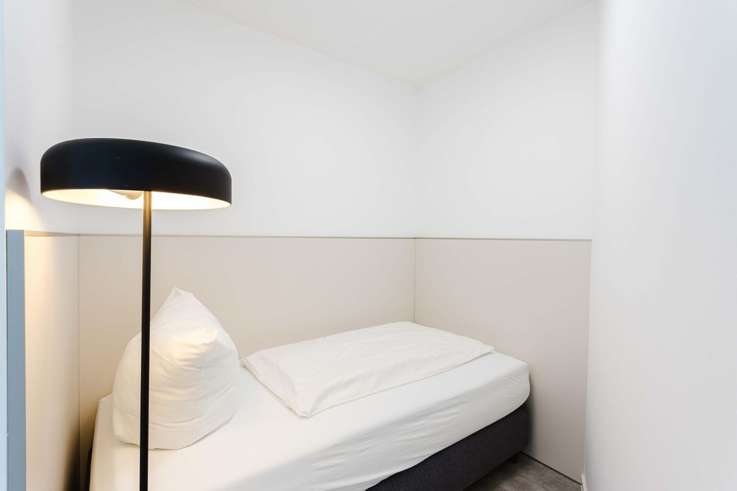 Un petit lit bien fait, avec une literie blanche et une lampe de sol noire, dans une pièce au décor minimaliste.
