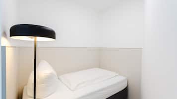 Ein kleines, ordentlich bezogenes Bett mit weißer Bettwäsche und einer schwarzen Stehlampe in einem minimalistisch eingerichteten Raum.
