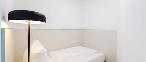 Una cama pequeña y ordenada con ropa blanca y una lámpara de pie negra en una habitación minimalista.