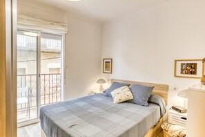 A bedroom with a bed, a nightstand, a lamp, a framed picture, and a balcony with a chair.