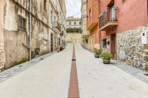 A narrow street with buildings on either side, featuring balconies with potted plants, a central drainage grate, and a stone wall on the right.
