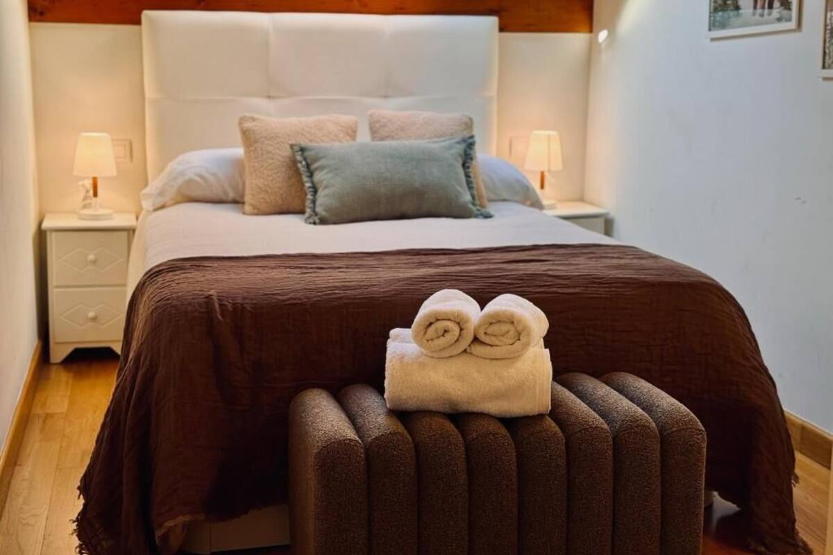 Una cama con una manta marrón, dos almohadas y una toalla enrollada sobre un banco de madera.