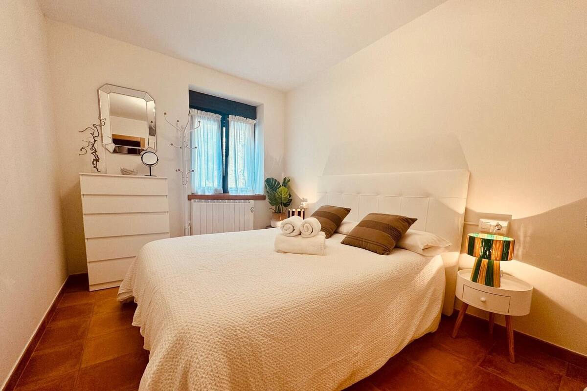 Un dormitorio con cama, cómoda, mesita de noche, lámpara y ventana con cortinas.