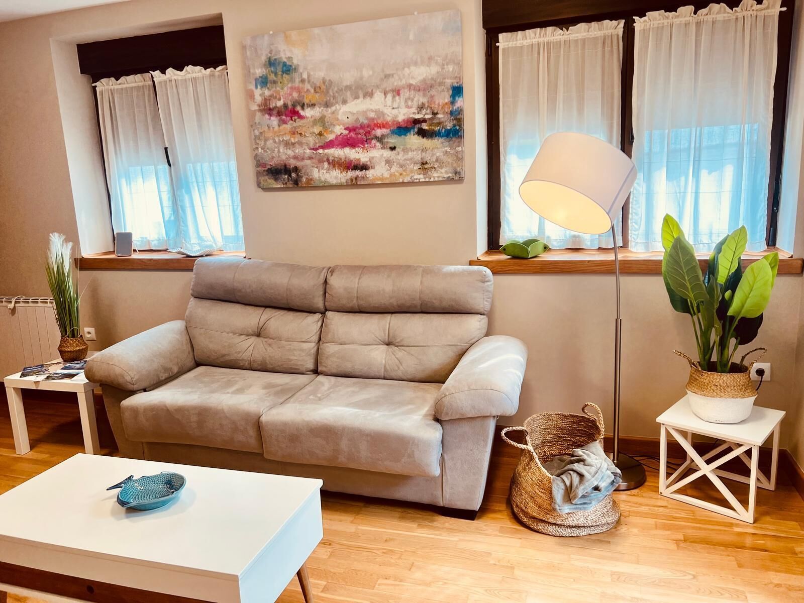 A living room with a beige sofa, a white coffee table, a floor lamp, a painting, and a window with curtains.