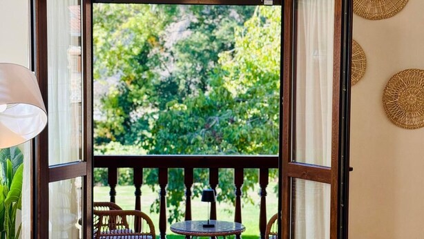 Un balcón abierto con vistas a la vegetación, una silla de mimbre, una mesita con una lámpara y un sofá.