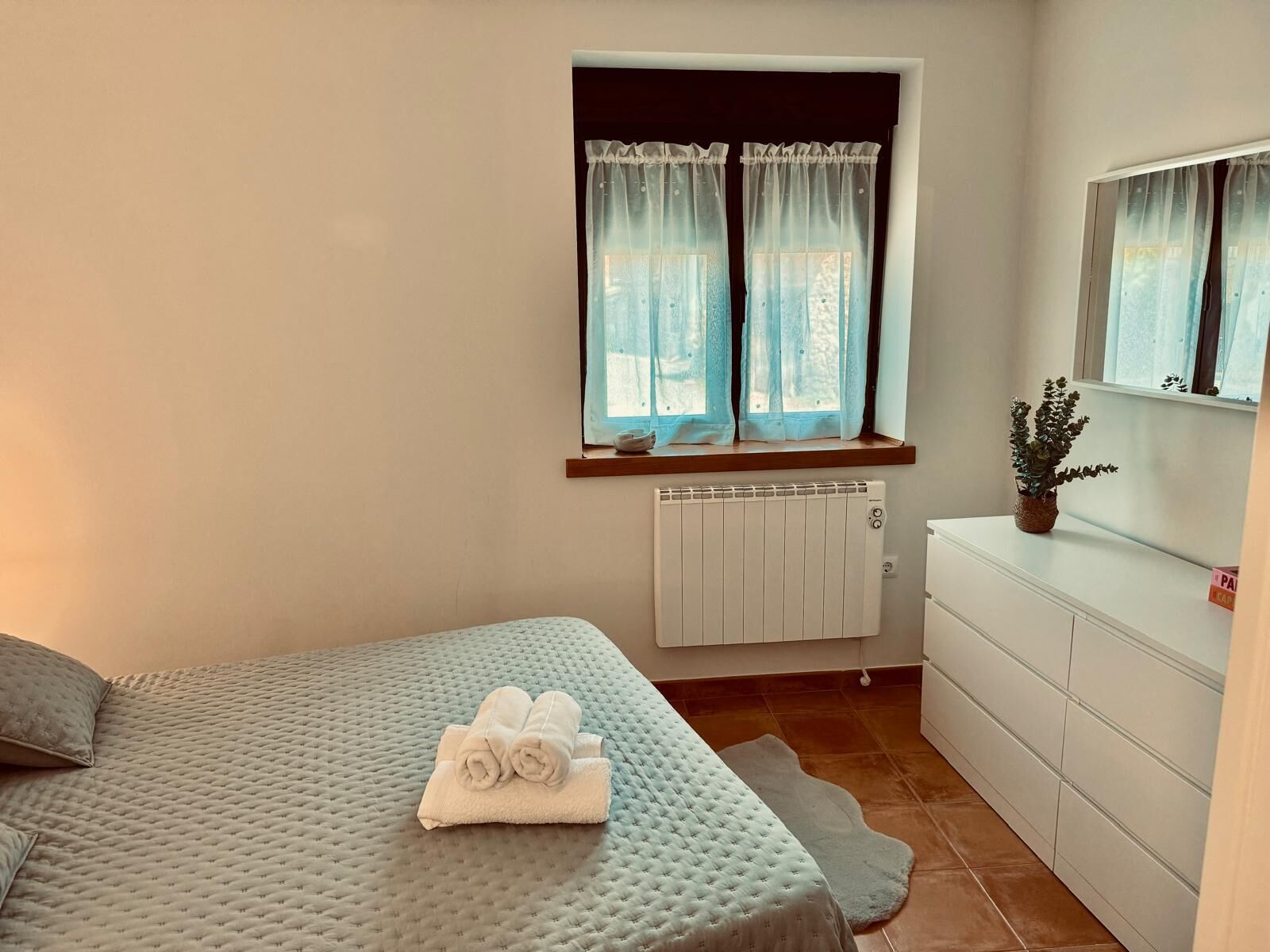 A bedroom with a bed, a radiator, a window with curtains, a dresser, and a potted plant.