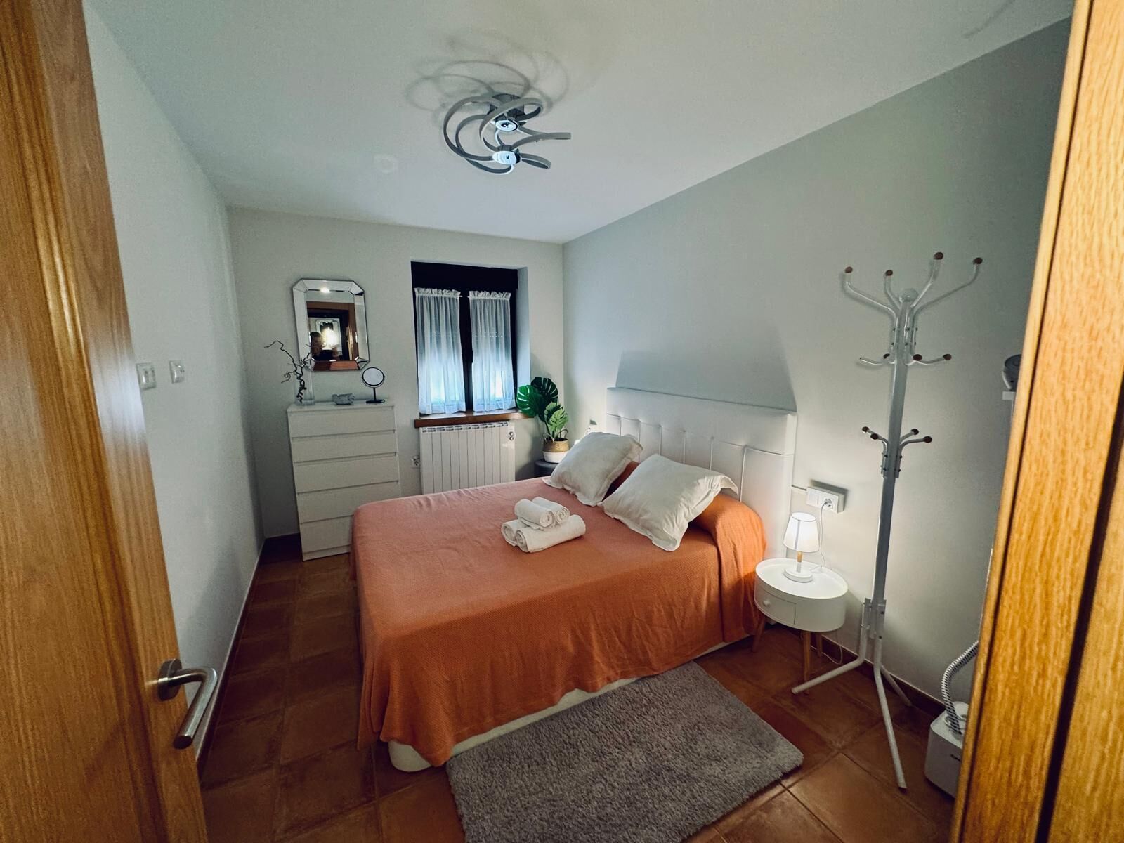 Un dormitorio con cama, cómoda, espejo, perchero y ventana con cortinas.